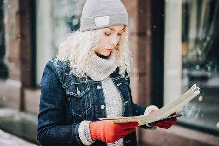 Young blond curly female tourist in warm clothes and red gloves with London map thoughtfully looking for a way in winter city, blurred facade on the backgroundの写真素材