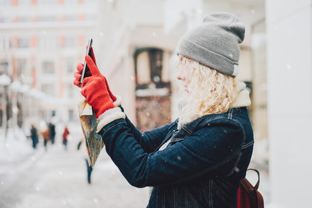 Young blond curly female tourist in warm clothes and red gloves with London map making photo or selfie on smartphone, winter city, blurred winter backgroundの写真素材