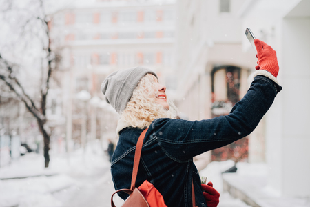 Young blond curly female tourist in warm clothes and red gloves with London map making selfie, winter city, blurred backgroundの写真素材