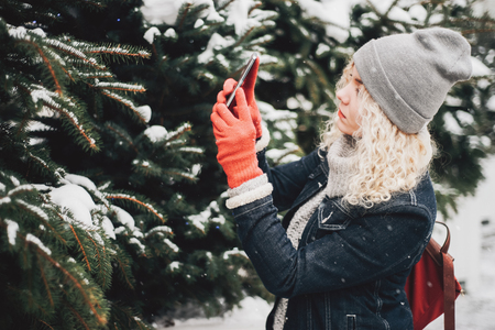 Young blond curly female tourist in warm clothes, red gloves and bag making photo of spruce on smartphoneの写真素材