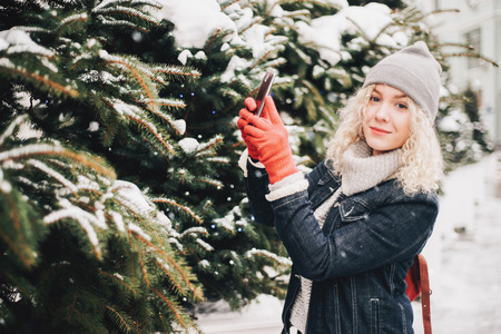 Young blond curly female tourist in warm clothes and red gloves making photo of spruce or selfie on a smartphoneの写真素材