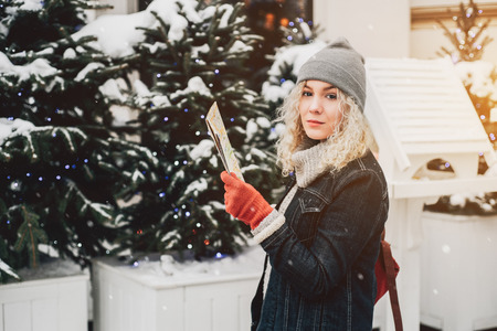 Young blond curly female tourist in warm clothes and red gloves with London map surrounded by snow-covered trees in winter backgroundの写真素材