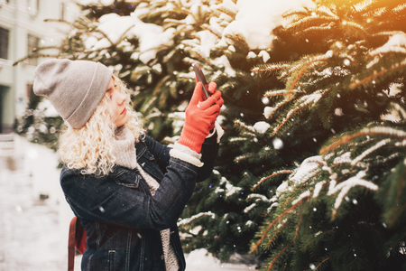 Young blond curly female tourist in warm clothes and red gloves making photo of spruce on a smartphoneの写真素材