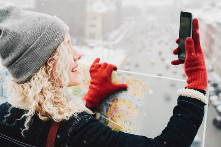 Young blond curly female tourist in warm clothes and red gloves with London map making selfie, standing on the roof of winter city, blurred road with cars in backgroundの写真素材