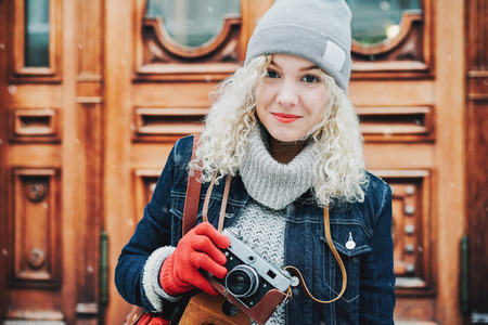 Young blond curly female tourist in warm clothes with old film camera and city map standing on the background of wooden doors, winter, snowの写真素材
