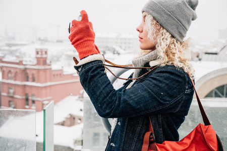 Young blond curly smiling female in warm clothes, red gloves and bag standing on the roof and shooting on retro film camera, background of winter city, snowの写真素材