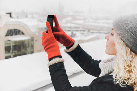 Young blond curly female tourist in warm clothes and red gloves making selfie or photo, ? standing on the roof of winter cityの写真素材