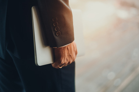View of hand of a married business man in formal dark elegant business suit holding white laptop, standing next to window of high floor, blurred sunny city highway in backgroundの写真素材