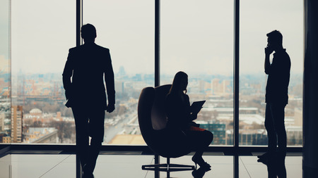Silhouettes of business people: businessman with businesswoman waiting for their third colleague which is coming to start meeting, high floor, winter cityscape outside, luxury office interiorの写真素材