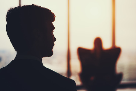 Silhouette of bearded businessman in formal suit standing in front of window and his female colleague who sitting on armchair in office interior, blurred cityscape with sunset outsideの写真素材