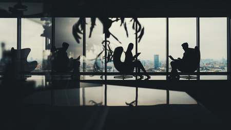Silhouettes of two business people man and woman working on their gadgets sitting on armchairs in office interior with a lot of reflections, with huge windows and winter cityscape outsideの写真素材