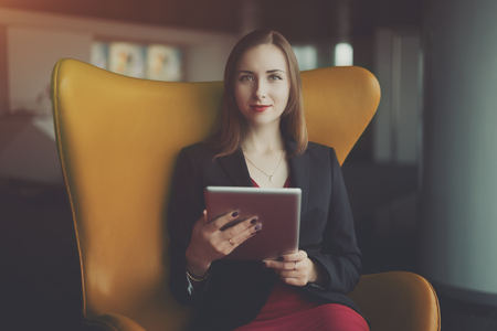 Portrait of young successful woman entrepreneur in a red dress and jacket sitting on orange armchair and working on her digital tablet, office interiorの写真素材