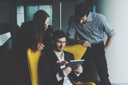 Group of young business people during meeting: man and woman standing next to yellow armchair with their boss in formal suit with digital tablet, discussing something and looking on screen of tabletの写真素材