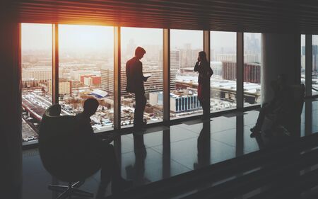 Businessman and businesswoman standing near window of skyscraper, two their male colleagues sitting on armchairs on opposite sides, winter cityscape outside, office interior with reflectionsの写真素材
