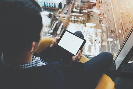 Mock up template of tablet pc in hands of businessman sitting near window of skyscraper and using modern digital tablet with blank screen for your text message, winter cityscape and highway outsideの写真素材