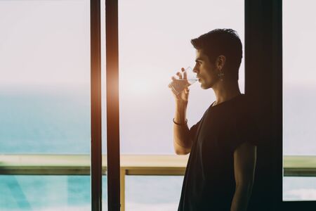 Handsome brazilian guy with earring, drinking water from transparent glass on the balcony near window on sunset, blue blurred ocean and horizon outside, Rio de Janeiro, Brazilの写真素材