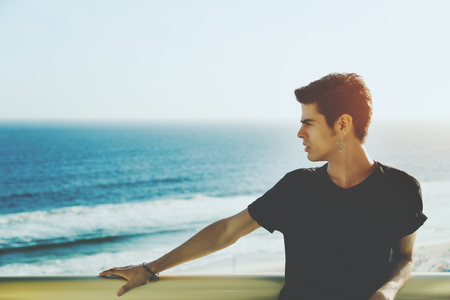 Handsome brazilian guy with earring standing on the balcony on sunny summer day, blue blurred ocean and horizon in background, Rio de Janeiro, Brazilの写真素材