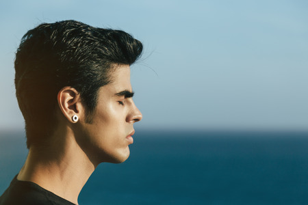 Portrait of young handsome brazilian guy with black hair and earring standing in front of ocean with eyes closed, breathing fresh sea air, sunny summer day, clear sky, Rio de Janeiroの写真素材