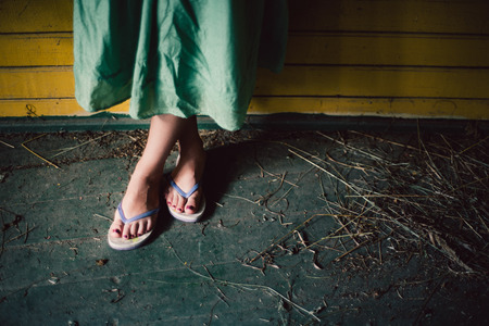 View of female feet in opened slippers with painted nails and greenish dress, yellow wooden wall on the background, dry grass or hay aroundの写真素材