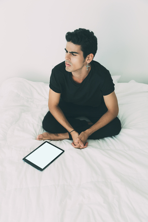 Handsome pensive brazilian guy with cross-earring in black t-shirt and tights is sitting on bed near mock up of digital tablet with white screen, in the morning after sleeping, white bedroom aroundの写真素材