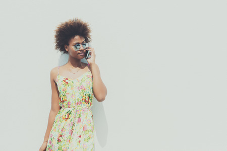 Black beautiful attractive young brazilian girl with curly hair in sunglasses and dress, talking on smartphone in front of white textured wall on sunny summer dayの写真素材