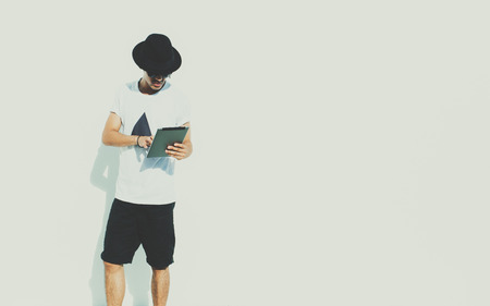 Attractive handsome young modern brazilian guy in sunglasses, white t-shirt, black hat and shorts using digital tablet, standing in front of textured white wall, sunny bright summer dayの写真素材