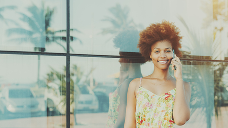 Black beautiful attractive young brazilian teen girl with curly hair in dress, smiling while talking on smartphone standing in front of tiled glass wall with reflections of palms and cars, sunny dayの写真素材