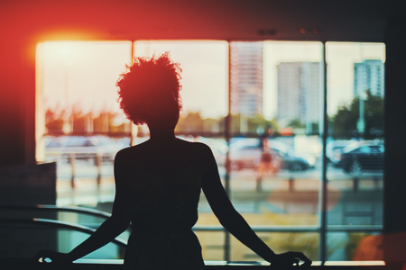 Silhouette of young black curly Brazilian girl in front of window standing in supermarket with blurred escalator behind her and cityscape out of the window, Rio de Janeiro, Brazilの写真素材
