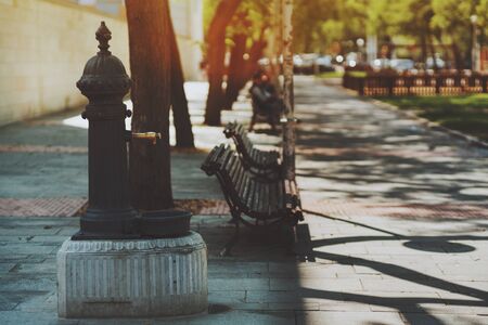 Vintage rusty brass or bronze water tap  standing on concrete base in park of Barcelona with several maroon benches near walking pathway of paving stone with shadows from treesの写真素材
