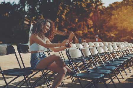 Curly brunette smiling beautiful girl on empty seat rows of folding chairs in park before concert making selfie on her digital tablet, attractive young woman using her touch pad in park before eventの写真素材