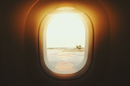 Contrast view through airplane porthole of take-off field, airport staff, fuel trucks; illuminator of airplane with landscape outside on sunny summer dayの写真素材