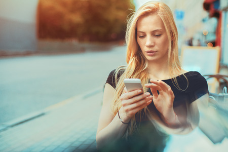 Pensive beautiful blonde caucasian girl sitting in street bar and texting message to her friend, thoughtful freelancer woman browsing site on her smart phone in cafe with copy space for your messageの写真素材