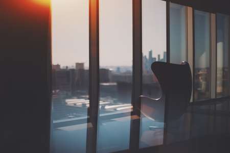 Silhouette of empty curved armchair standing on reflective floor of modern office next to window with cityscape outside and silhouettes of Moscow City business skyscrapers group on sunny dayの写真素材