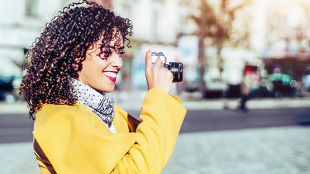 Beautiful laughing curly Brazilian woman in yellow coat is shooting on vintage camera, portrait of adult white smiling lady on sunny day using retro photo camera with copy space for your text messageの写真素材