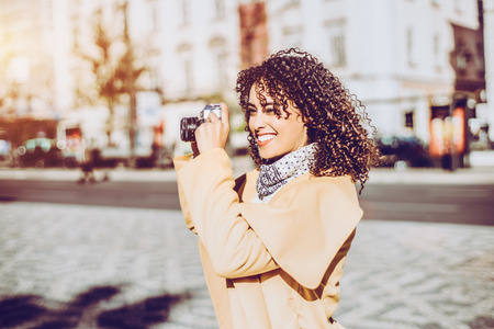 Smiling white curly woman in coat and sweater on sunny street with retro camera, young laughing Brazilian brunette with perfect smile shooting on vintage photo camera, blurred city road in backgroundの写真素材