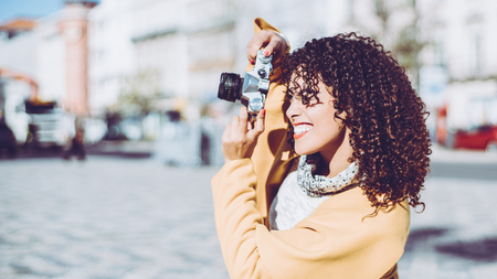 Beautiful curly Brazilian woman in yellow coat is making shoot on old vintage camera, portrait of adult white smiling lady on sunny day using retro photo camera with copy space for your text messageの写真素材