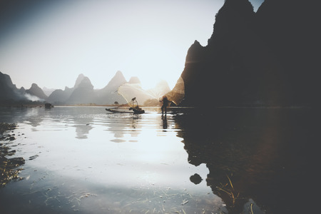 Wide angle shoot of silhouette of cormorant fisherman in China throwing a net into the river while standing on ancient traditional Chinese bamboo boat at sunrise with mountains and hills in backgroundの写真素材