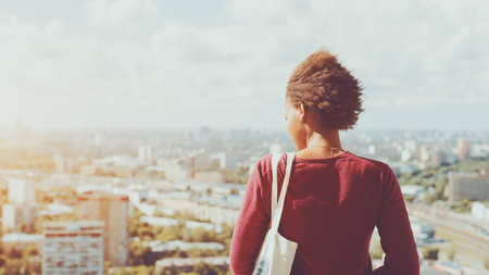 Rear view of young mixed Brazilian teen girl with curly afro hair waving in the wind standing on high point and looking down on city district, sunny summer cityscape below in blurred backgroundの写真素材