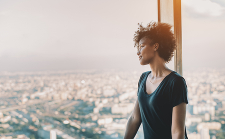 Attractive young mixed girl in black shirt and with curly african hair is standing near window of very high tower observation point and pensively looking on evening cityscape of modern metropolitanの写真素材