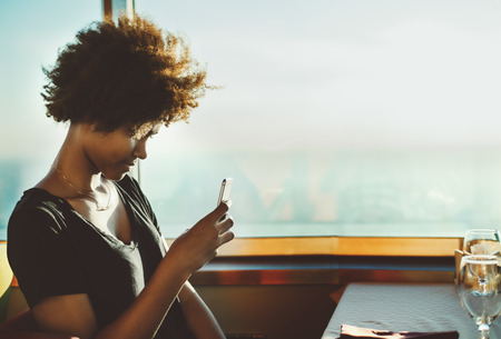 Pensive biracial girl is using her smart phone to make selfie while sitting in caffe near big window and waiting her food order, curly teenage hipster female with gadget in restaurant or cruise shipの写真素材