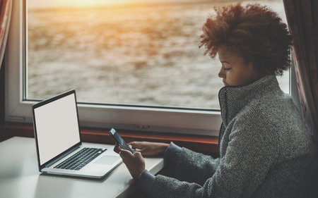 Serious curly female biracial teenager travelling by ship, sitting near window and sending message to her family while working on freelance job with modern laptop with white blank screen mock-upの写真素材