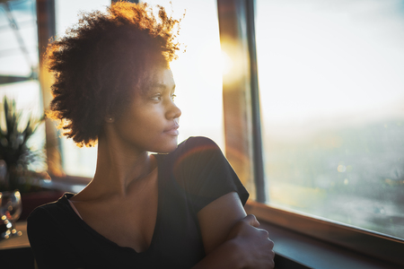 Curly charming young afro american tourist female in black dress is sitting in luxury restaurant of cruise ship near window and thoughtfully looking out the big window on ocean and warm sunset horizonの写真素材