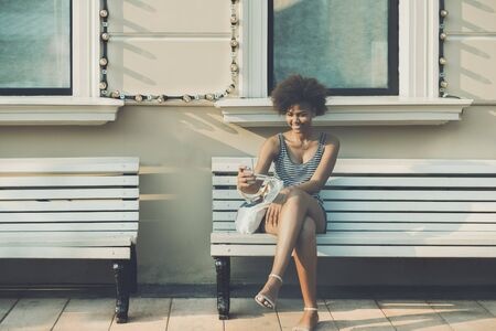 Cheerful curly Brazilian girl making selfie while sitting on bench during summer day withs copy space for text, charming laughing teenage female with smart phone surfing her social network accountsの写真素材
