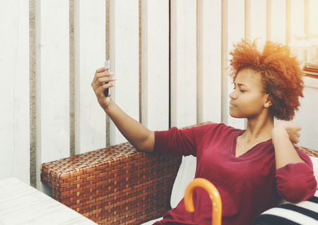 Teenage curly black female is sitting in street restaurant and making selfie while waiting her food, young mixed girl surfing her social network feed on smartphone while sitting on the chair outdoorsの写真素材