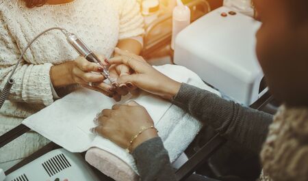 Close-up view of hands of afro american brown woman in nail salon receiving manicure, master preparing nails for shellac manicure using special rotating nail file machine deviceの写真素材
