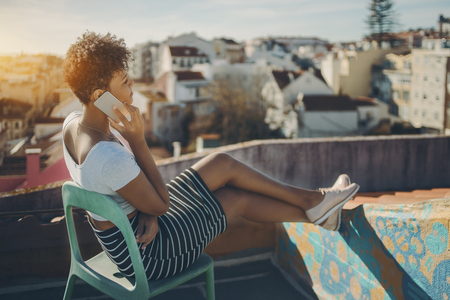 Charming afro american lady sitting on teal chair on roof of Lisbon residential house and talking via smart phone, young Brazilian girl having rest on balcony of European city and phoning with friendの写真素材