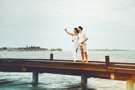 Beautiful newly married couple making selfie using modern smartphone on Maldives resort during their honeymoon while standing on bridge, with copy space zone for logo, text or advertising messageの写真素材