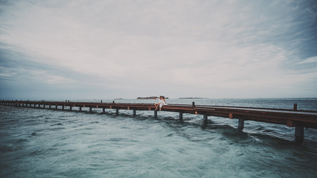 Adult caucasian couple man and woman are making selfie using modern smartphone on Maldives resort while sitting in the centre of bridge on warm windy weather with overcast sky and waves on ocean waterの写真素材