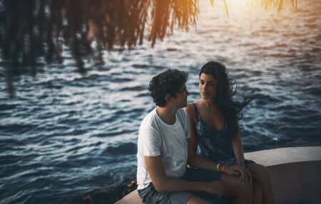 Adult couple: Brazilian man and Spanish woman sitting under sunshade on ocean pier and holding each others hands with ocean water in blurred background, Maldives resort on sunny eveningの写真素材