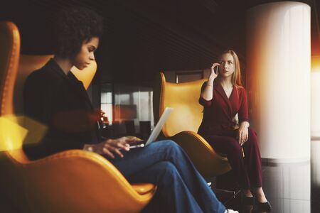 Two businesswoman sitting in luxury office interior on yellow curved armchairs: black young trainee female with modern laptop and adult caucasian woman talking by smartphone with her bossの写真素材